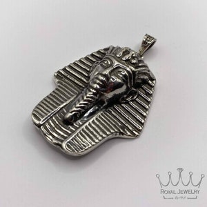 May include: Silver pendant featuring a detailed depiction of an Egyptian pharaoh's head, with a textured design resembling a sarcophagus.