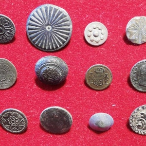 Könnte beinhalten: Sortiment von zehn Vintage-Metallknöpfen in verschiedenen Formen, Größen und Designs. Einige Knöpfe haben ein Sternburst-Muster, andere ein Blumenmuster und einige ein Wappen. Die Knöpfe sind in Silber-, Gold- und Schwarztönen.