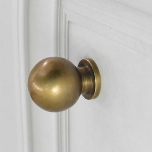 Peut inclure: Un bouton de meuble sphérique de couleur laiton, monté sur une porte blanche. Le bouton a une forme de boule arrondie avec une base circulaire. La quincaillerie est d'une teinte métallique chaude, contrastant avec la porte blanche.
