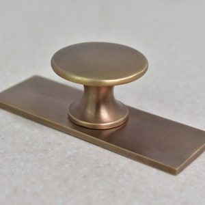 May include: A brushed brass cabinet knob with a rectangular base plate. The knob has a round top and a slightly flared base.