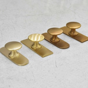May include: Four brass cabinet knobs with a rounded top and a base plate. The knobs are arranged in a row from left to right, with the base plates facing the viewer. The knobs are different shades of brass, ranging from a light gold to a darker bronze.