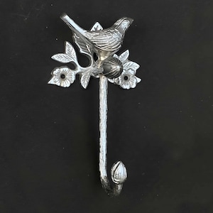 May include: Silver metal hook with a bird perched on a branch with leaves and flowers. The hook is shaped like a branch and has a rounded tip.