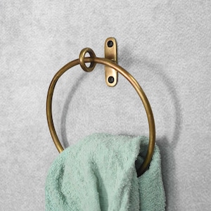 May include: A brass towel ring with a simple design. The ring is mounted to a wall with a single screw. A light green towel is hanging on the ring.