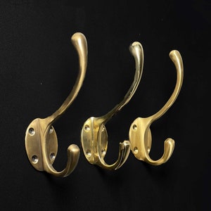 May include: Three gold-toned metal hooks mounted on a black background. The hooks are curved and have a simple design.