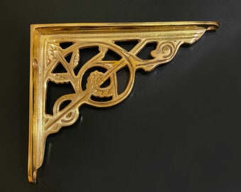 Op de afbeelding: Een gouden metalen plankbeugel met een decoratief, versierd ontwerp. De beugel heeft een bloemmotief en is rechthoekig gevormd.