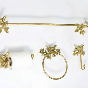 Op de afbeelding: Gouden badkamerset met een handdoekrek, toiletpapierhouder, handdoekring en haak. Elk onderdeel heeft een decoratief vogeltje dat op een tak met bladeren zit.