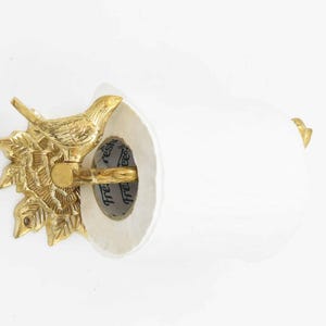 Op de afbeelding: Gouden metalen toiletpapierhouder met een vogelontwerp. De houder is bevestigd aan een witte muur. De houder heeft een witte papierrol met de tekst "fresh" en "reshi" erop.