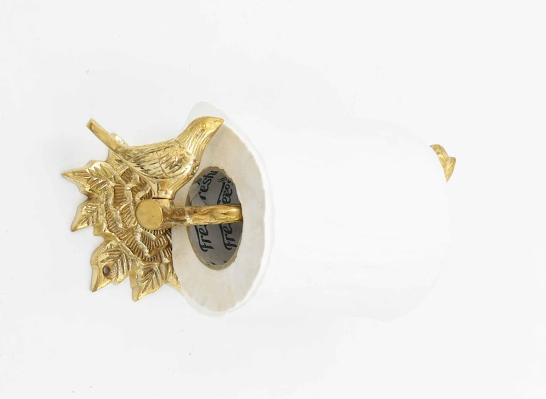 Op de afbeelding: Gouden metalen toiletpapierhouder met een vogelontwerp. De houder is bevestigd aan een witte muur. De houder heeft een witte papierrol met de tekst "fresh" en "reshi" erop.