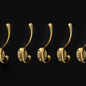 May include: Five gold-toned metal hooks with a rounded base and a double hook design. The hooks are mounted on a black background.