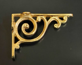 Op de afbeelding: Een gouden metalen plankbeugel met een decoratief scrollontwerp. De beugel heeft een grote spiraal en kleinere scrollen aan de randen.