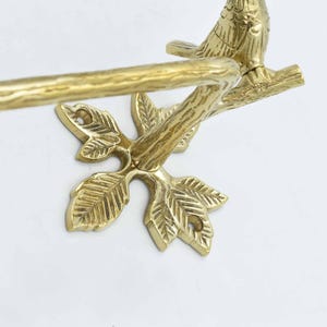 Op de afbeelding: Gouden metalen handdoekrek met een ontwerp van een vogel op een tak. Het rek heeft een bladvormige basis.