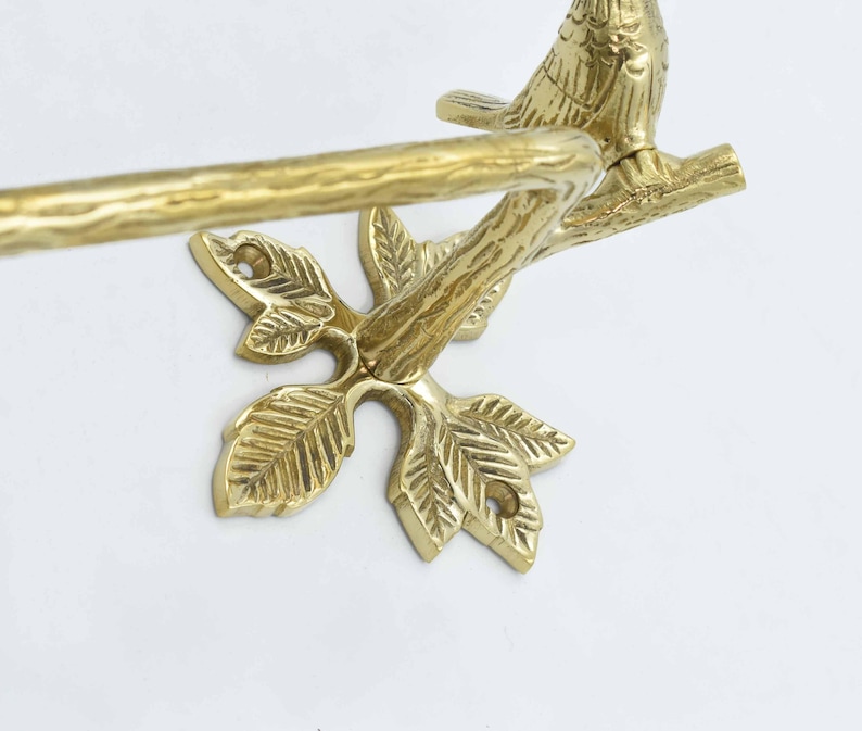 Op de afbeelding: Gouden metalen handdoekrek met een ontwerp van een vogel op een tak. Het rek heeft een bladvormige basis.