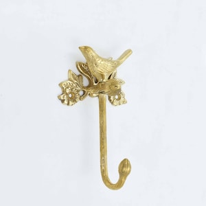 Op de afbeelding: Gouden metalen haak met een vogel die op een tak zit met bladeren en bloemen. De haak is ontworpen om aan de muur te worden gemonteerd.