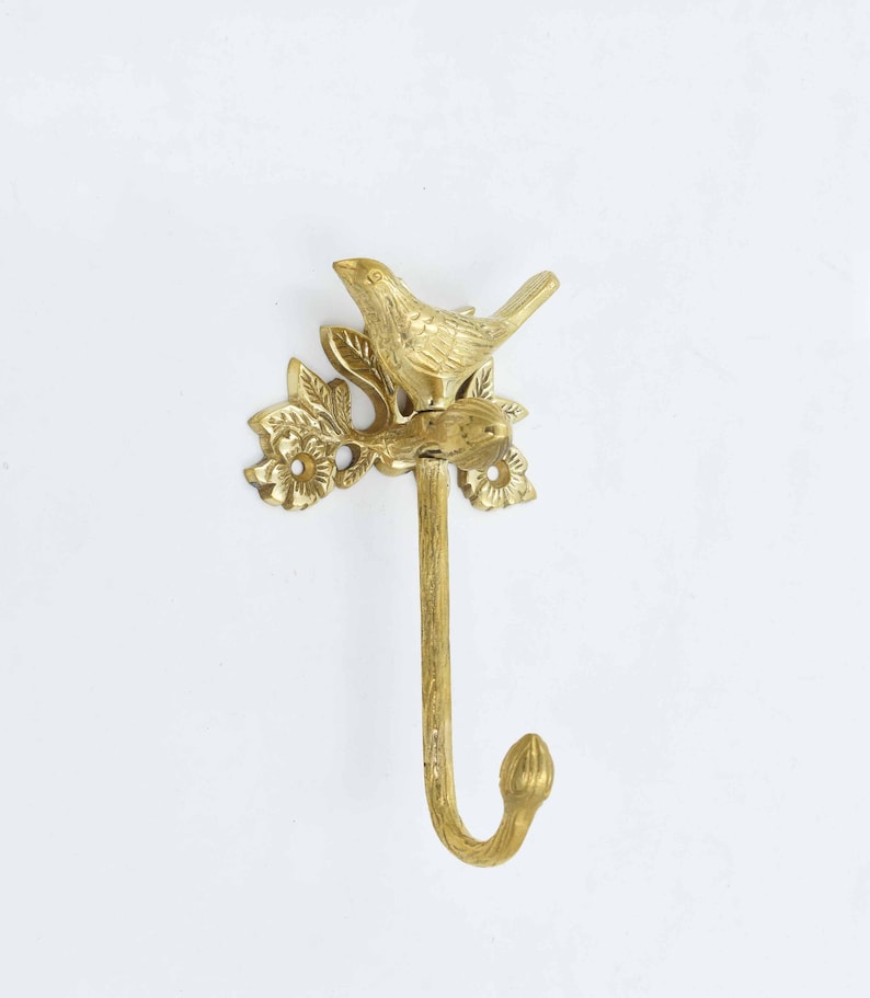 Op de afbeelding: Gouden metalen haak met een vogel die op een tak zit met bladeren en bloemen. De haak is ontworpen om aan de muur te worden gemonteerd.