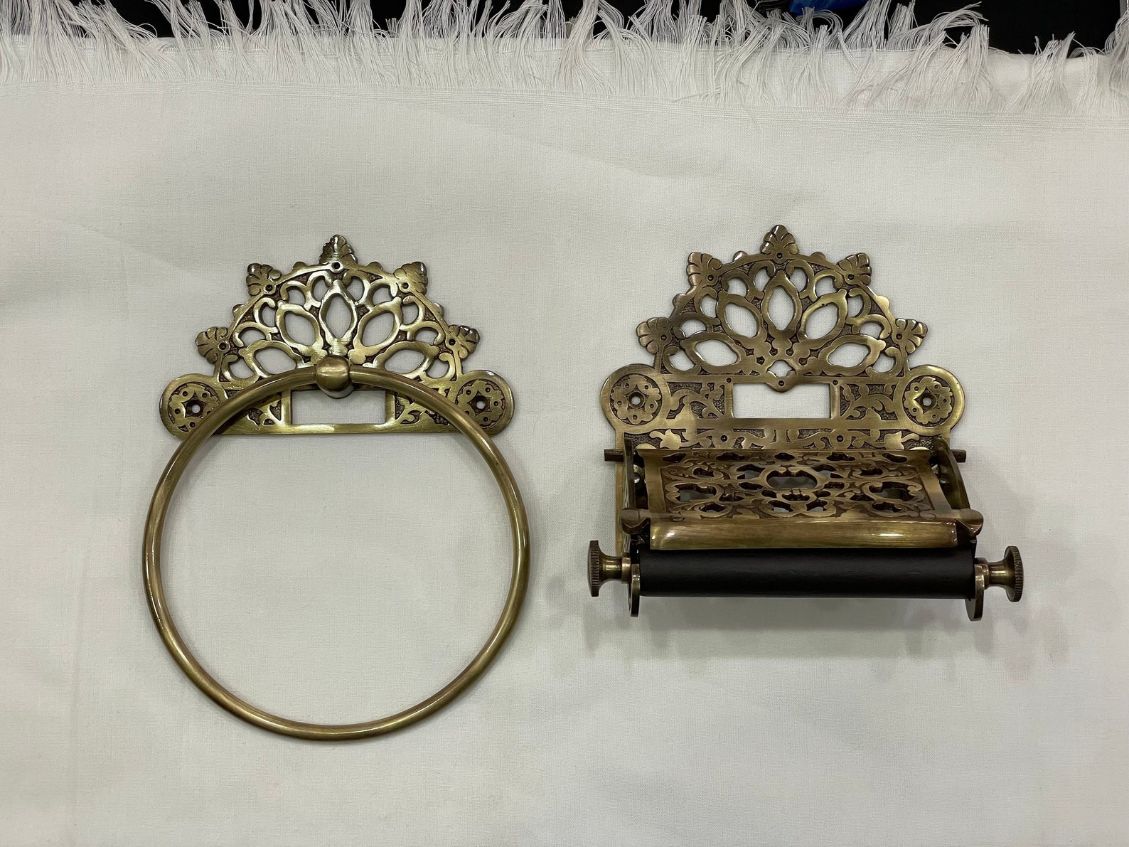 Ornate brass towel ring and toilet paper holder with floral scrollwork, displayed on white fabric