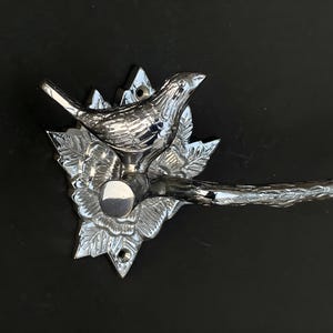 May include: Chrome-plated metal toilet paper holder with a bird and floral design. The holder has a curved arm and a decorative base.