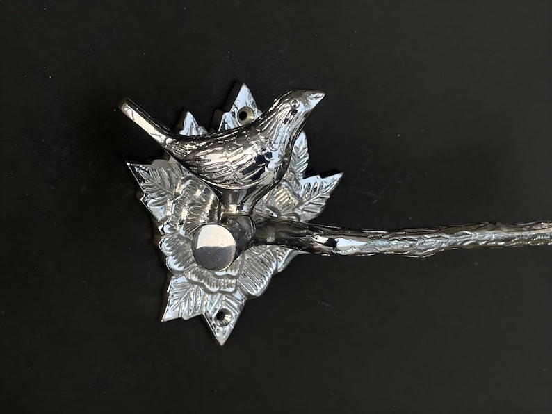 May include: Chrome-plated metal toilet paper holder with a bird and floral design. The holder has a curved arm and a decorative base.
