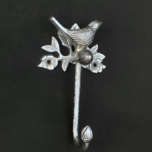 May include: Silver metal hook with a bird perched on a branch with leaves and flowers. The hook is shaped like a tree trunk and has a bud at the bottom.