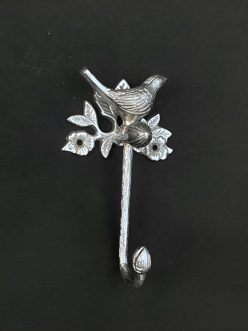 May include: Silver metal hook with a bird perched on a branch with leaves and flowers. The hook is shaped like a tree trunk and has a bud at the bottom.