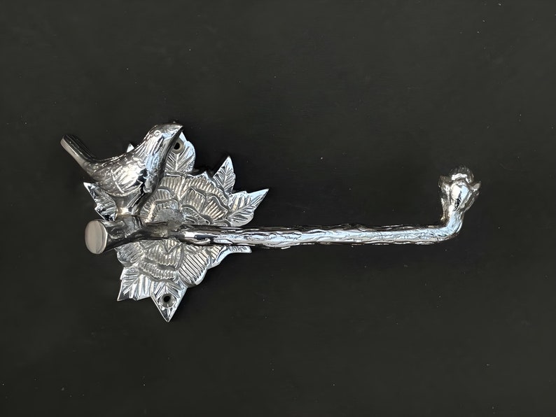 May include: Silver-toned metal toilet paper holder with a bird and floral design. The holder has a branch-like arm and a decorative base with a bird perched on a branch.