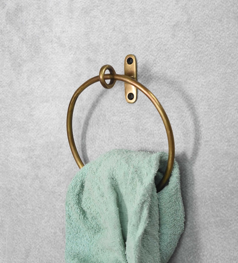 May include: A brass towel ring with a simple design. The ring is mounted to a wall with a single screw. A light green towel is hanging on the ring.