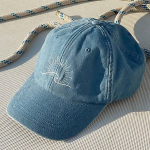 Può includere: Un berretto da baseball in denim blu con un sole e un'onda ricamate in bianco.