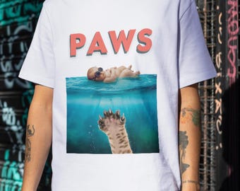 May include: White t-shirt with a graphic of a baby wearing goggles floating in blue water with a cat paw reaching up from below. The text "PAWS" is printed in red above the graphic.