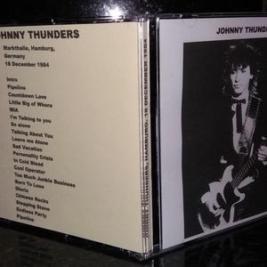 Puede incluir: Una caja de CD con Johnny Thunders. El lado izquierdo muestra texto, incluida la ubicación y la fecha del concierto. El lado derecho tiene una imagen en blanco y negro de Johnny Thunders tocando la guitarra. La caja del CD está en una funda de plástico transparente.
