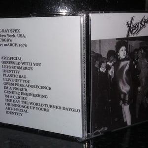 May include: A CD case featuring the band X-Ray Spex. The cover includes a black and white photo of the band performing. The case also displays the band's name, location, and a list of song titles.