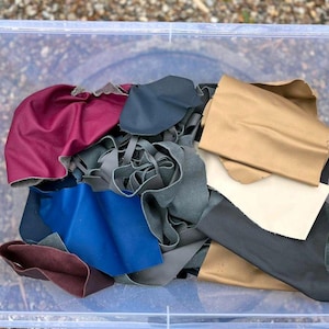 Könnte beinhalten: Eine transparente Plastikbox gefüllt mit Lederresten in den Farben Burgunderrot, Blau, Beige und Grau. Die Lederstücke haben unterschiedliche Formen und Größen, was auf eine Sammlung zum Basteln oder für Upcycling hindeutet.