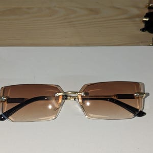 Puede incluir: Gafas de sol sin montura con lentes degradadas marrones y montura de metal dorado. Las lentes rectangulares están conectadas por un fino puente dorado y presentan pequeños detalles decorativos. Las patillas son negras.