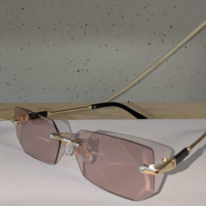 Può includere: Occhiali da sole senza montatura con montatura color oro e lenti rosa sfumate. Le aste sono dorate con dettagli neri vicino ai terminali. Le lenti hanno una forma geometrica.