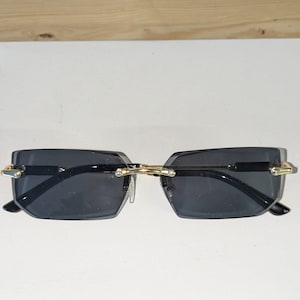 May include: Rimless rectangular sunglasses with dark lenses and gold-coloured metal detailing. The sunglasses feature a bridge connecting the lenses and thin, gold-coloured temples. The glasses are set against a white background.