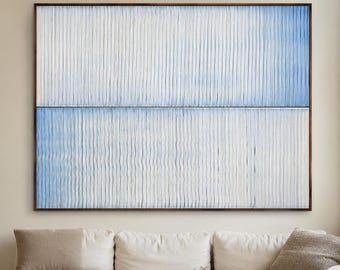 May include: Large abstract wall art with a textured white and blue design in a dark wood frame. The artwork hangs above a white sectional sofa and a rustic wooden coffee table in a neutral-toned living room.