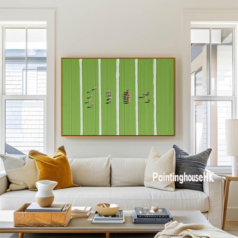 May include: A framed abstract painting of a green field with white lines and red and white figures. The painting is hanging on a white wall above a beige couch with yellow and gray pillows.