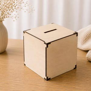 Peut inclure: Une boîte en bois de couleur claire avec une fente sur le dessus pour insérer des objets. La boîte est de forme carrée avec des bordures marron foncé sur les coins et les bords. La boîte est sur une surface en bois.