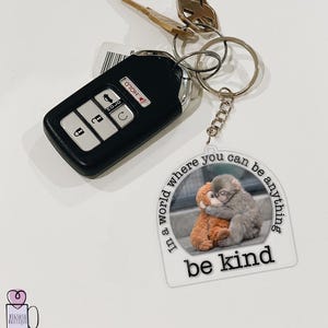 May include: A keychain with a clear, circular charm featuring a gray monkey hugging an orange stuffed animal. The charm has the text "In a world where you can be anything, be kind." Attached to the keychain are keys and a black car key fob.