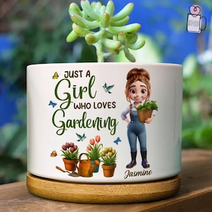 May include: A white ceramic planter with the text "Just a Girl Who Loves Gardening" and a cartoon illustration of a gardener. The planter holds a succulent plant and sits on a wooden base. Additional graphics include butterflies, flowers, and gardening tools.