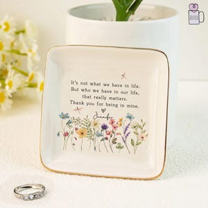 Può includere: Un vassoio quadrato in ceramica bianca con bordo dorato. Il vassoio presenta un motivo floreale e una citazione: "Non è ciò che abbiamo nella vita. Ma chi abbiamo nella nostra vita, che conta davvero." Anche il nome "Jennifer" è stampato.