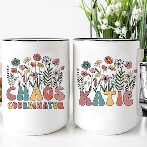 May include: Two white ceramic mugs with black handles. The mug on the left has a floral design and the text "Chaos Coordinator" in colorful letters. The mug on the right has a floral design and the text "Katie" in colorful letters.
