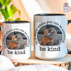 May include: Two white ceramic mugs with black handles and rims. Each mug features a circular design with a hugging monkey illustration and the text "In a world where you can be anything, be kind."