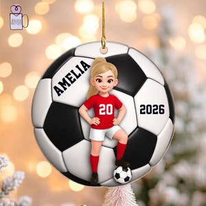 Può includere: Un ornamento rotondo in ceramica con un motivo a pallone da calcio bianco e nero. Un personaggio dei cartoni animati con capelli biondi e una maglia rossa con il numero 20 si trova sul pallone. Il nome AMELIA e l'anno 2026 sono stampati in nero.