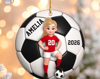 May include: A round ceramic ornament featuring a black and white soccer ball pattern. A cartoon character with blonde hair in a red jersey with the number 20 stands on the ball. The name AMELIA is printed in black, and the year 2026 is displayed.