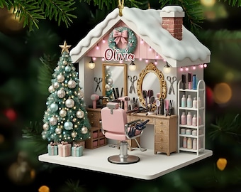 May include: A Christmas ornament shaped like a salon, featuring a pink barber chair, a vanity with a mirror, and a Christmas tree. The ornament has a white roof with snow and the name "Olivia" on the front.