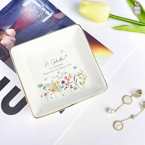 Pu&ograve; includere: Piatto quadrato in ceramica bianca con bordi dorati. Il piatto presenta il testo "A Godmother is a blessing. Thank you for being mine." e un motivo floreale. Il nome "Jennifer Mulber" e l'anno "2025" sono stampati sul piatto.
