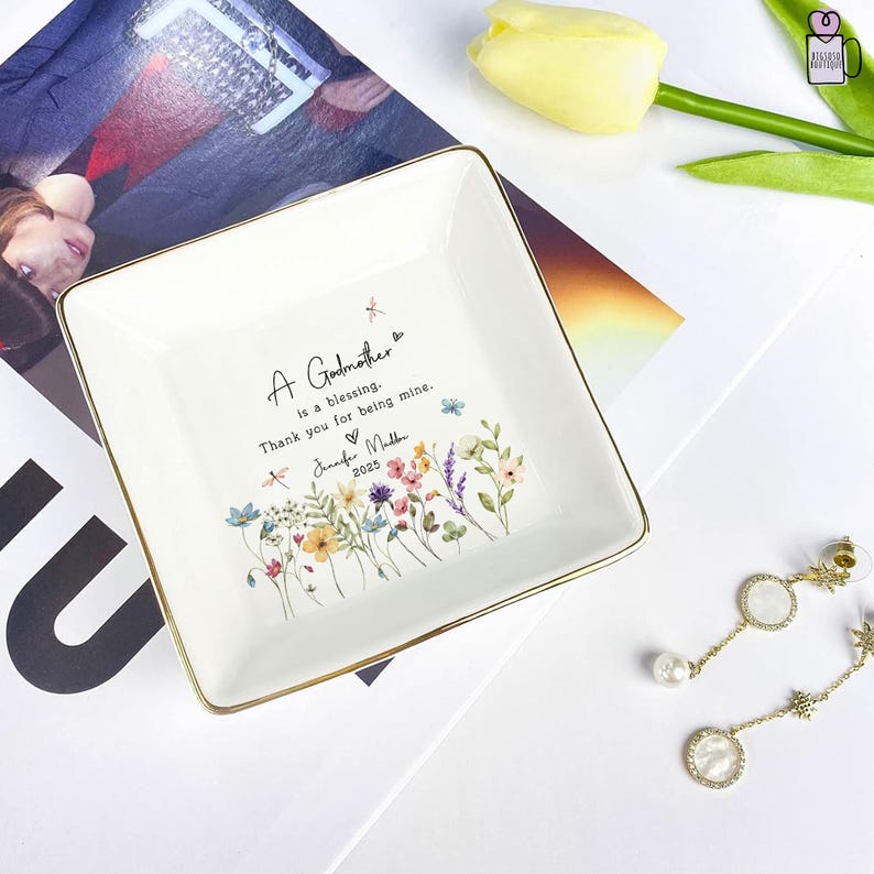 Pu&ograve; includere: Piatto quadrato in ceramica bianca con bordi dorati. Il piatto presenta il testo "A Godmother is a blessing. Thank you for being mine." e un motivo floreale. Il nome "Jennifer Mulber" e l'anno "2025" sono stampati sul piatto.