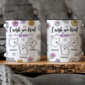 May include: Two white coffee mugs with black handles. The mugs have a floral design and a map of the United States with the text "I wish we lived closer" and the names Jessica, Ashley, and Patricia.