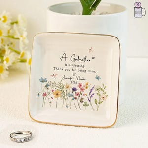 Pu&ograve; includere: Un vassoio quadrato in ceramica bianco sporco con bordo dorato, con opere d'arte floreali e la scritta "A Godmother is a blessing. Thank you for being mine." Include il nome "Jennifer Maddox" e l'anno "2025".