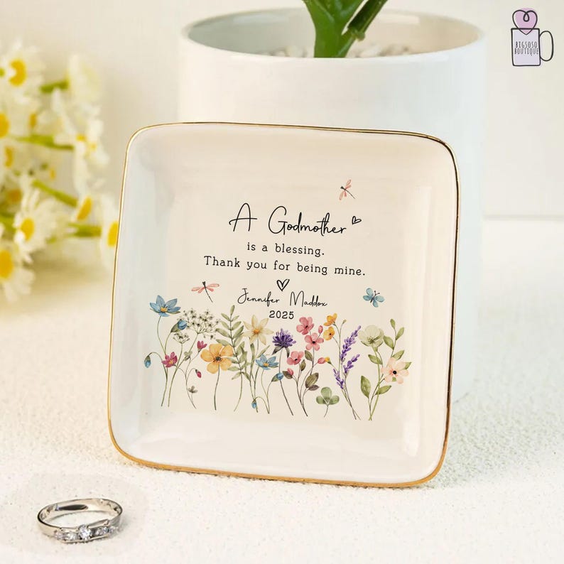 Pu&ograve; includere: Un vassoio quadrato in ceramica bianco sporco con bordo dorato, con opere d'arte floreali e la scritta "A Godmother is a blessing. Thank you for being mine." Include il nome "Jennifer Maddox" e l'anno "2025".