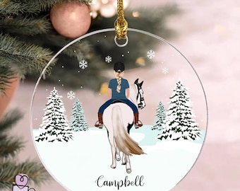 May include: A circular clear acrylic Christmas ornament featuring a printed illustration of a person with a blonde braid wearing a black cap and blue shirt riding a white horse through a snowy forest with pine trees, the name Campbell, and 2026.